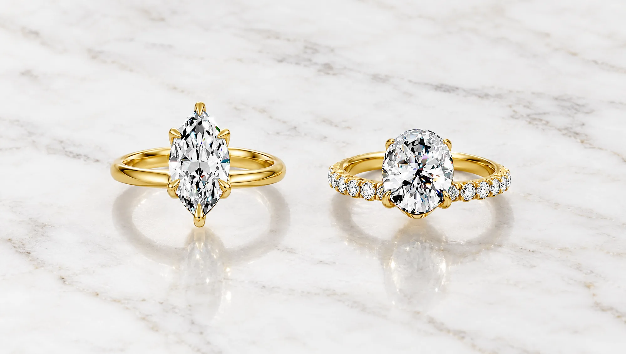 A yellow gold marquise diamond solitaire and a yellow gold oval diamond ring with a pavé band side by side on white marble