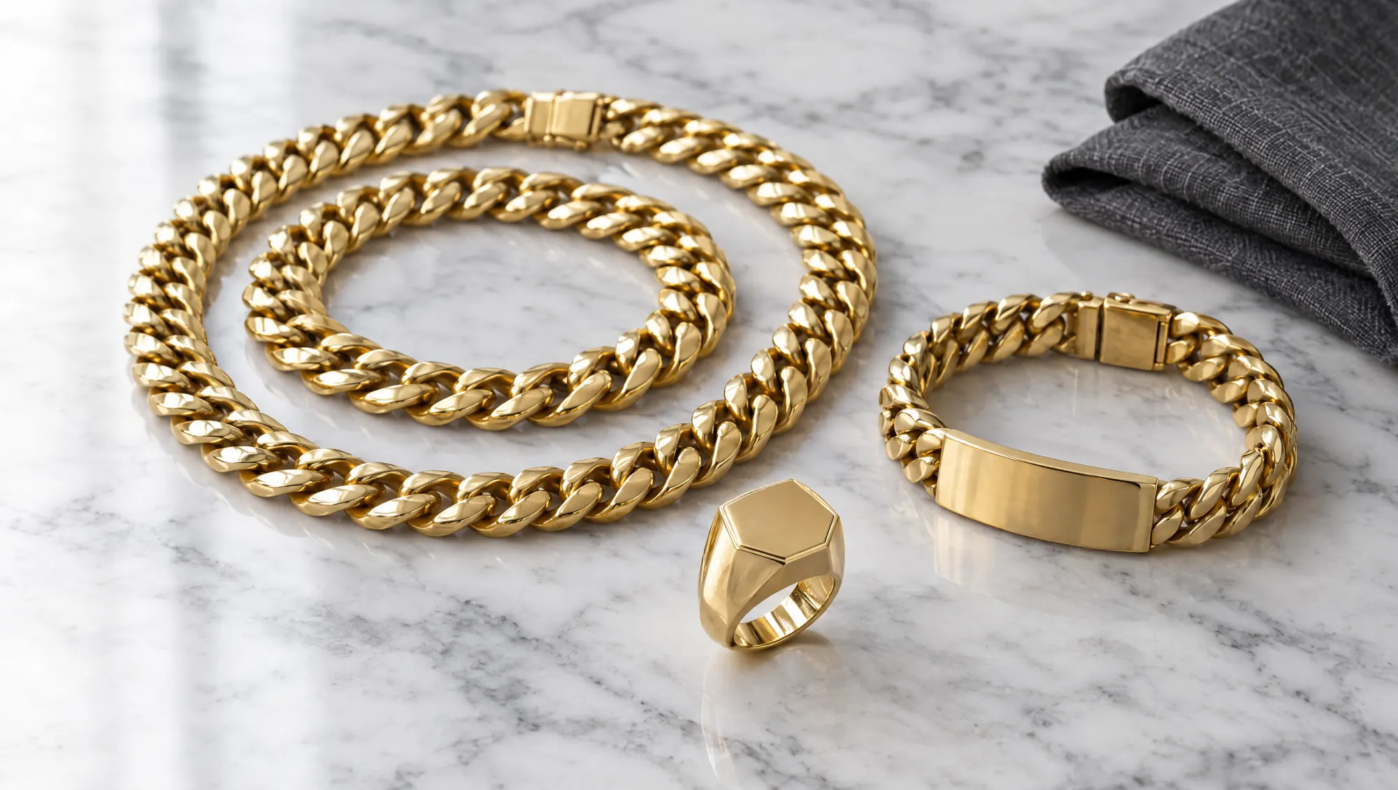 Gold Cuban link chain and bracelet, an ID-plate Cuban bracelet and a hexagonal signet ring on white marble with grey linen