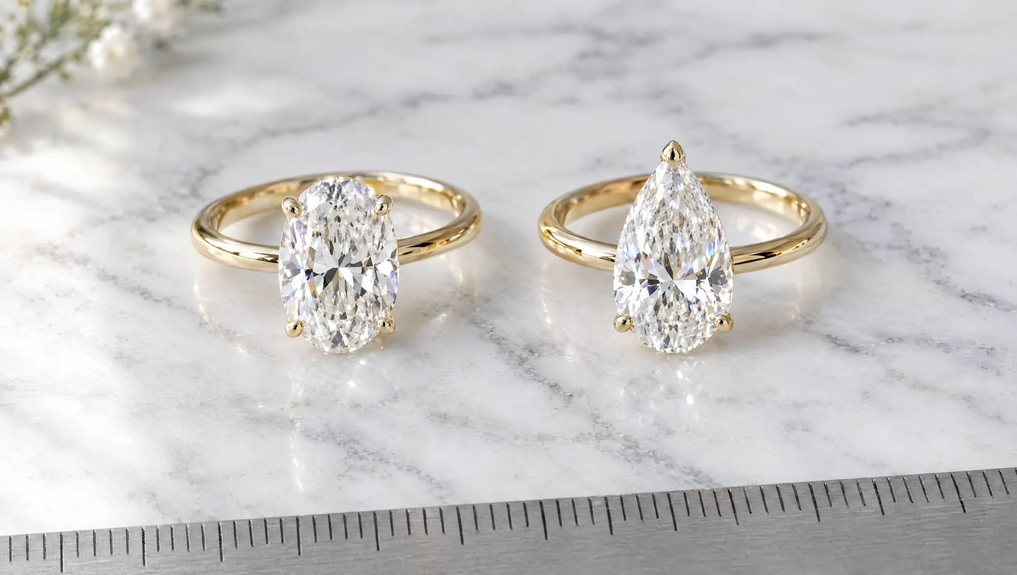 Oval and pear diamond solitaire rings in yellow gold side by side on white marble above a steel ruler