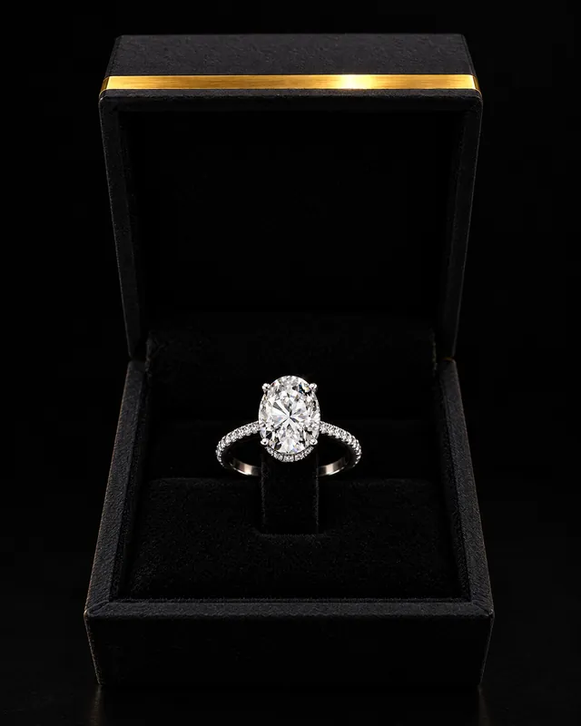 An oval diamond ring in an open black Mazimo ring box with a gold band on the lid